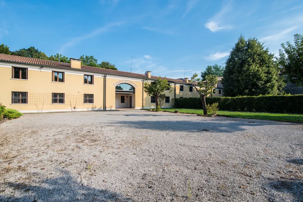 Exterior view of a yellow building with brown trim, a gravel courtyard, and green trees under a blue sky.