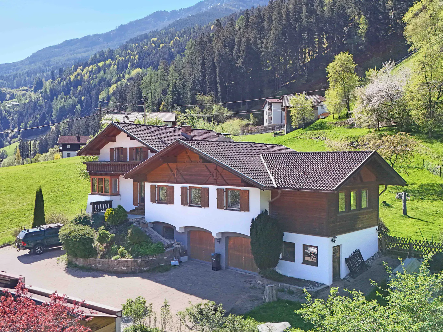 A white house with a brown roof and shutters sits on a green hillside with trees and mountains in the background.
