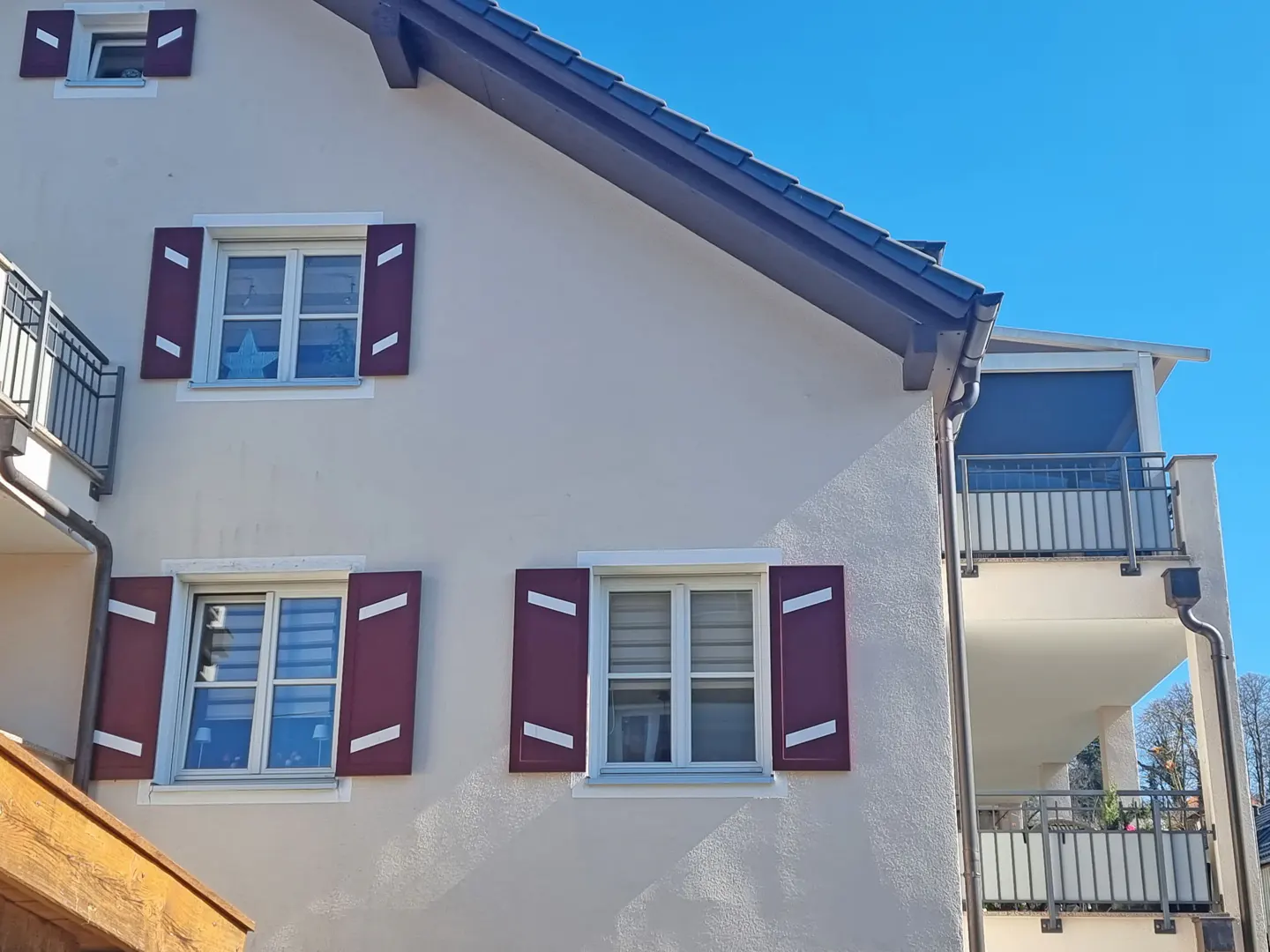 Exterior view of a light beige building with maroon shutters and a dark gray roof against a clear blue sky. Balconies with gray railings are visible.