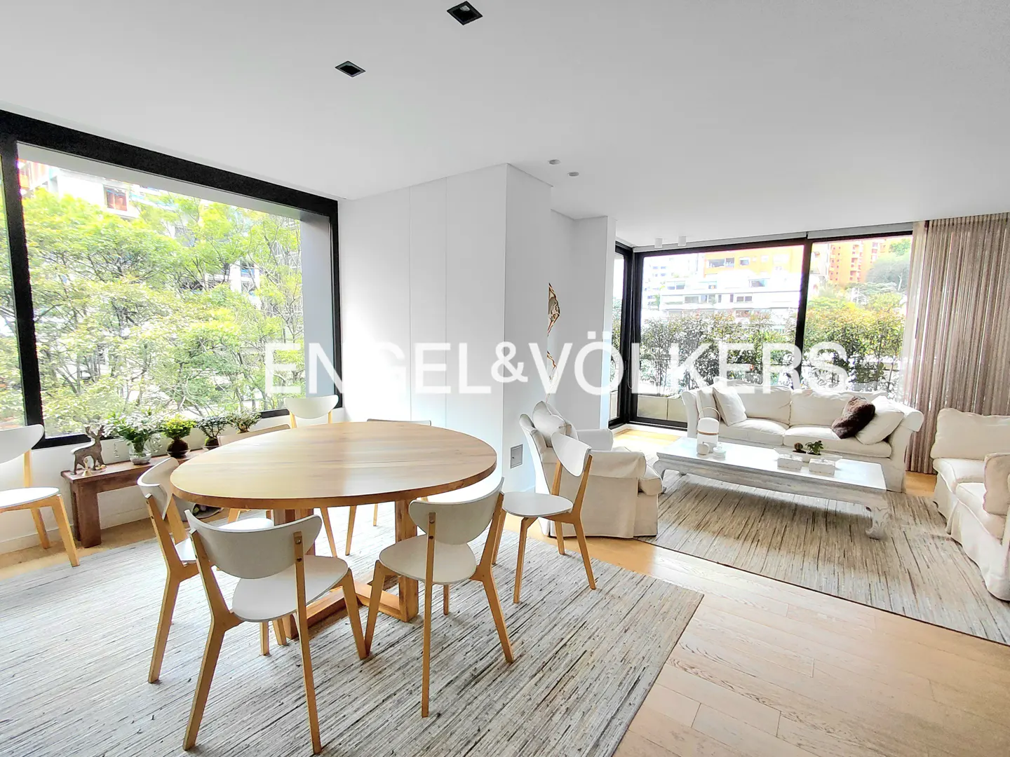 Bright, modern apartment interior with a wood table, white chairs, and a white sofa. Large windows overlook green trees.