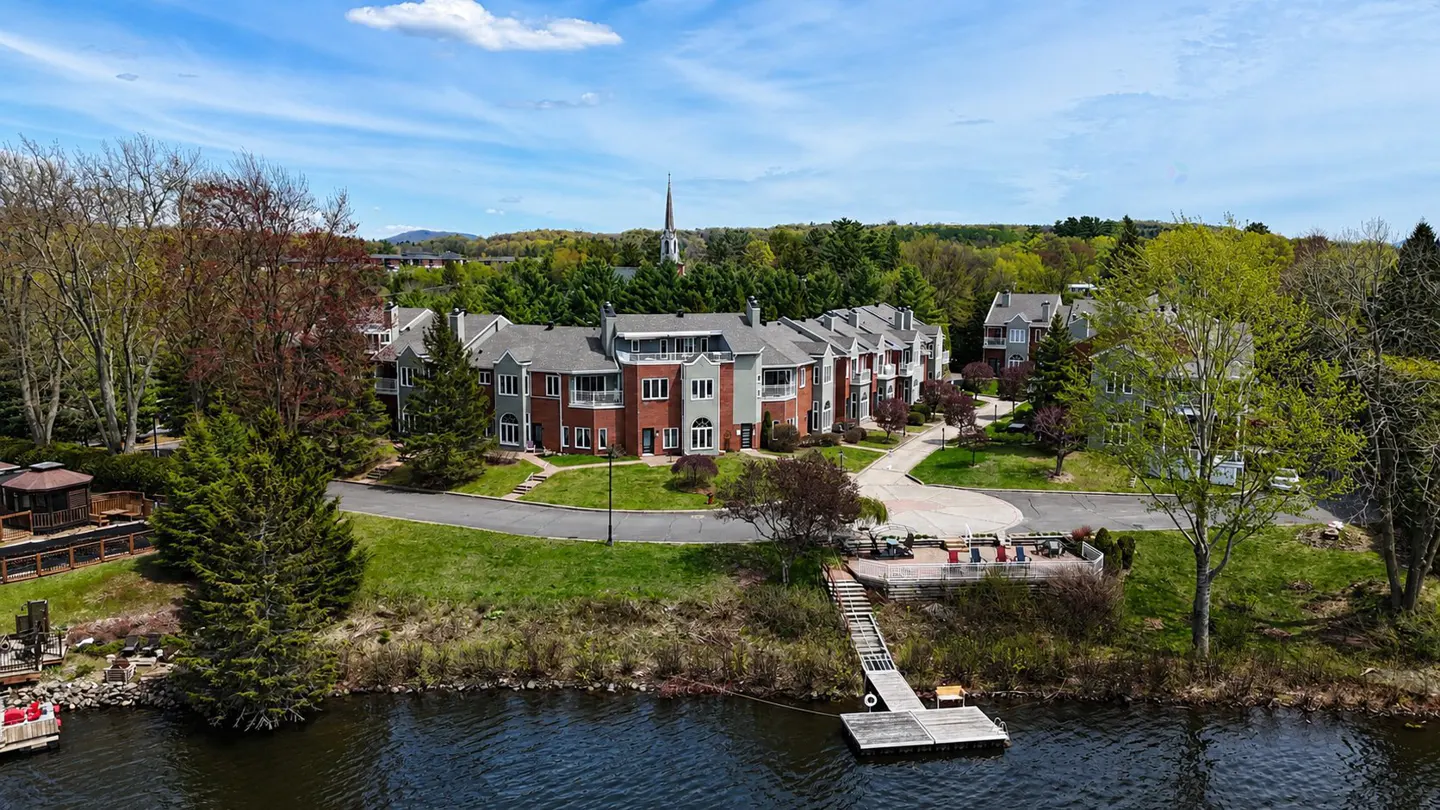 Aerial view of brick townhouses with balconies, a wooden dock on a lake, and a church steeple in the background.