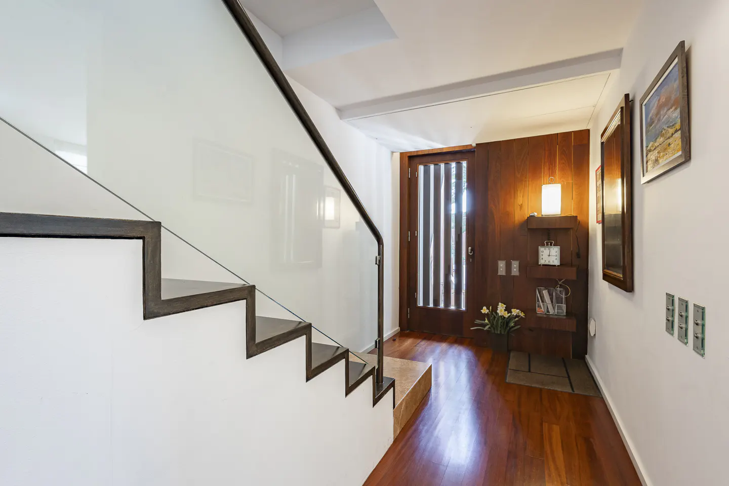 Interior view of a modern home entrance with a staircase featuring glass railings and a dark wood door with vertical slats. The walls are white, and the floor is polished wood.