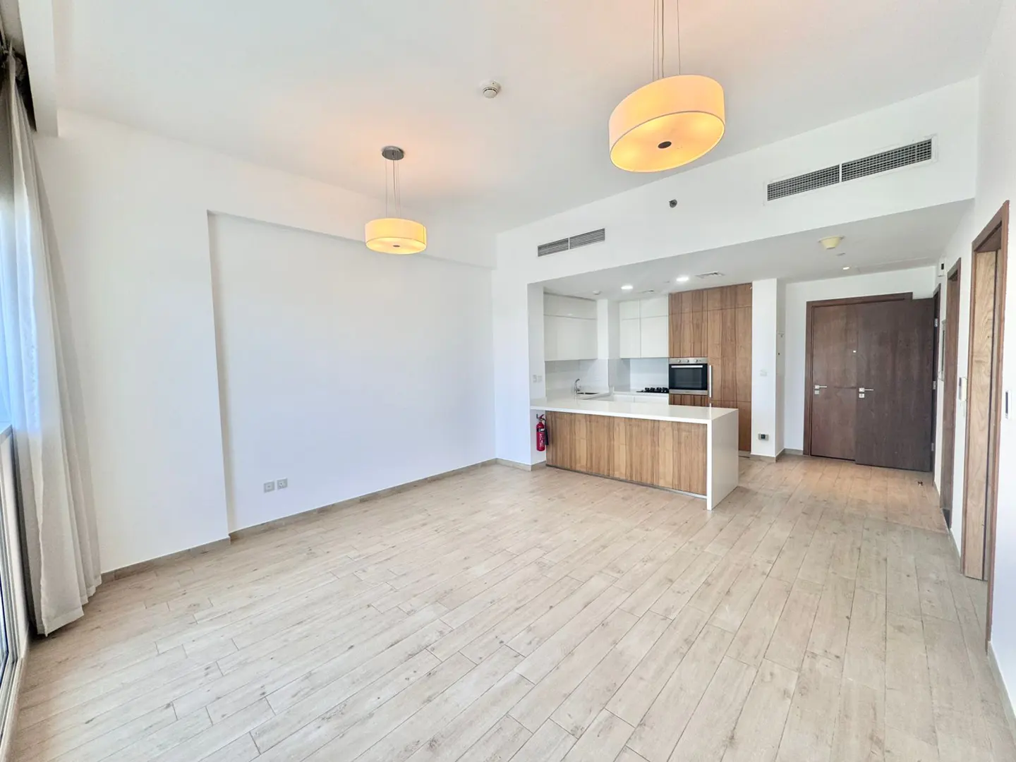 Bright, empty apartment with wood floors, white walls, and open kitchen. Two drum pendant lights hang from the ceiling.