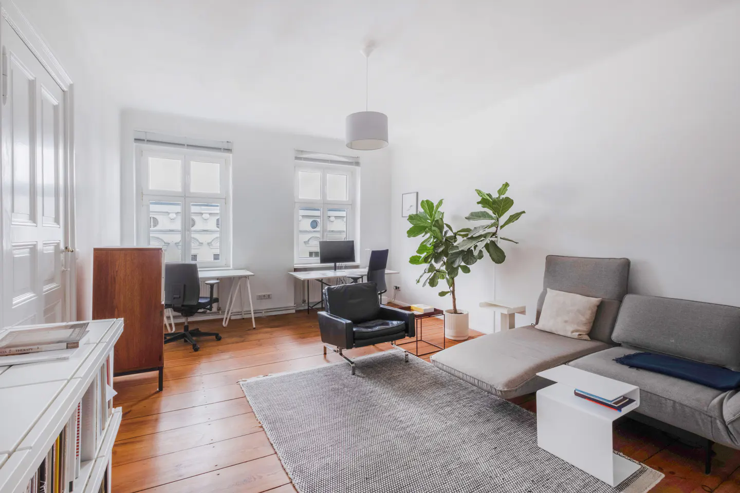 Bright, airy room with hardwood floors, white walls, desks, a gray sofa, and a large potted plant. Windows let in natural light.