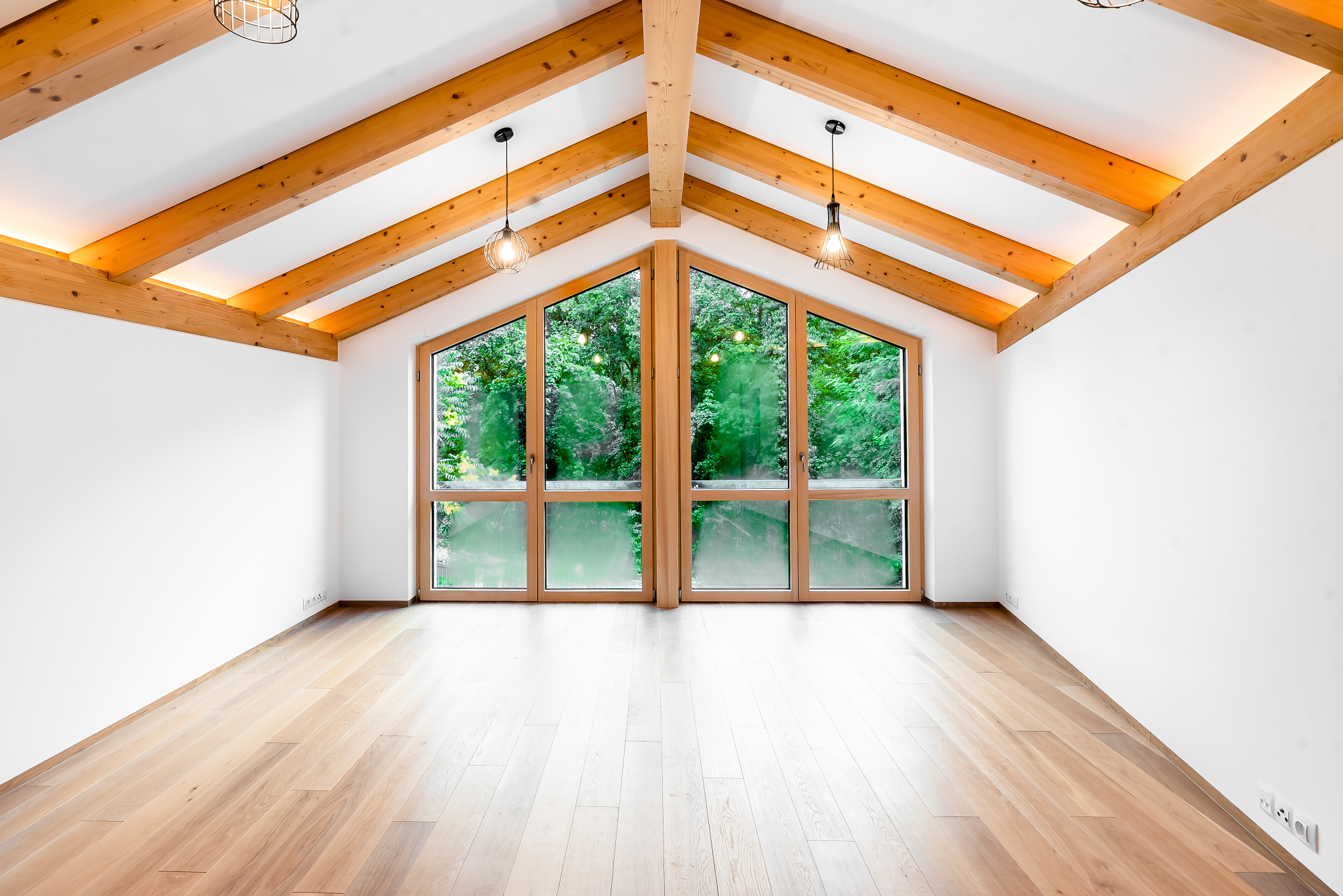 Empty room with light wood floors, white walls, exposed wooden ceiling beams, and large windows overlooking green trees. [AI-generated text]
