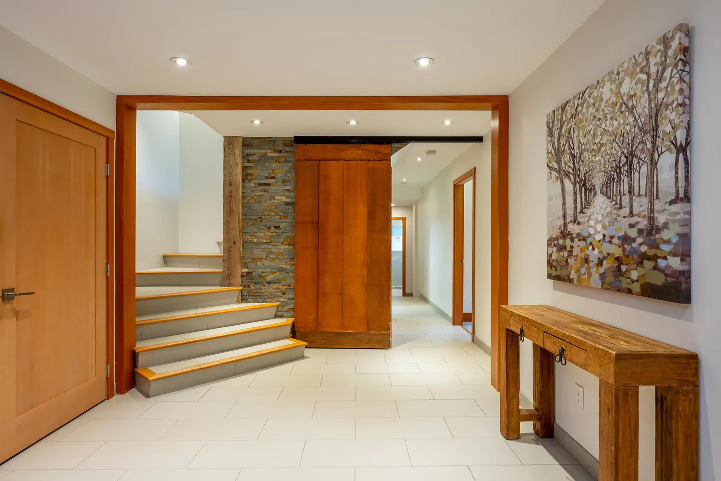 Hallway with a wooden sliding door, stone accent wall, stairs, and a rustic wooden console table with a tree painting above it.