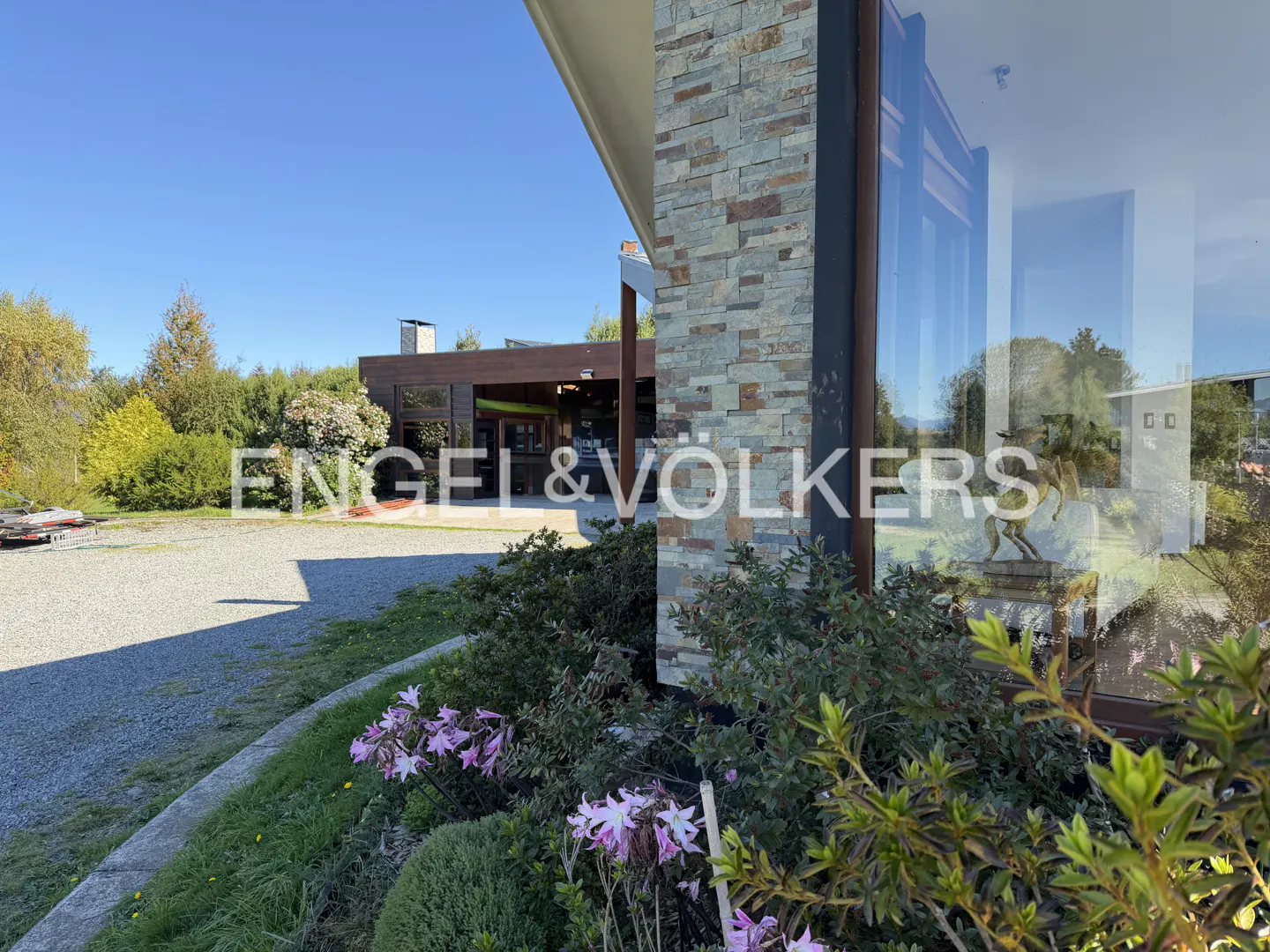 Exterior view of a modern home with stone accents, large windows, and a gravel driveway. Green foliage and flowers in the foreground. Blue sky.