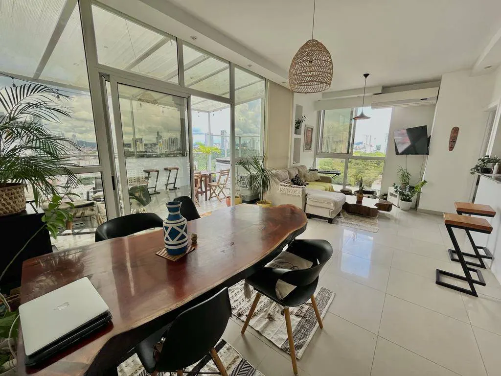 Bright, open-concept living space with a wood dining table, black chairs, and a laptop. Large windows offer city views. A woven pendant light hangs above.