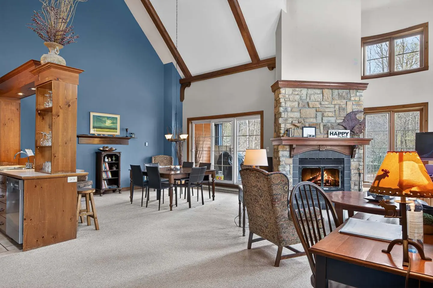 Open-concept living space with blue accent wall, stone fireplace, dining table, and wood beams.