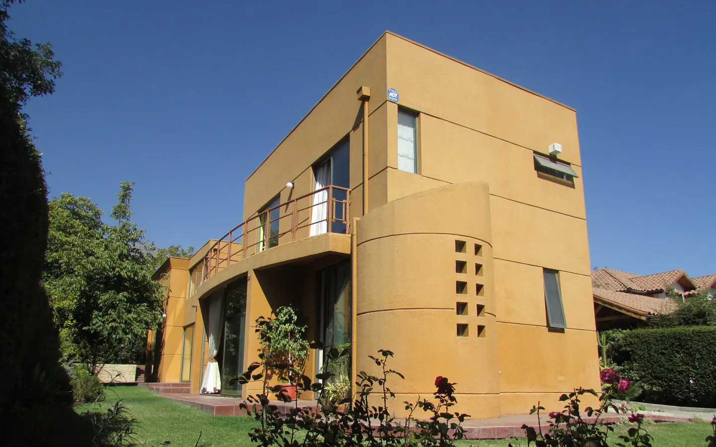 Two-story tan house with a curved balcony and a green lawn under a clear blue sky.