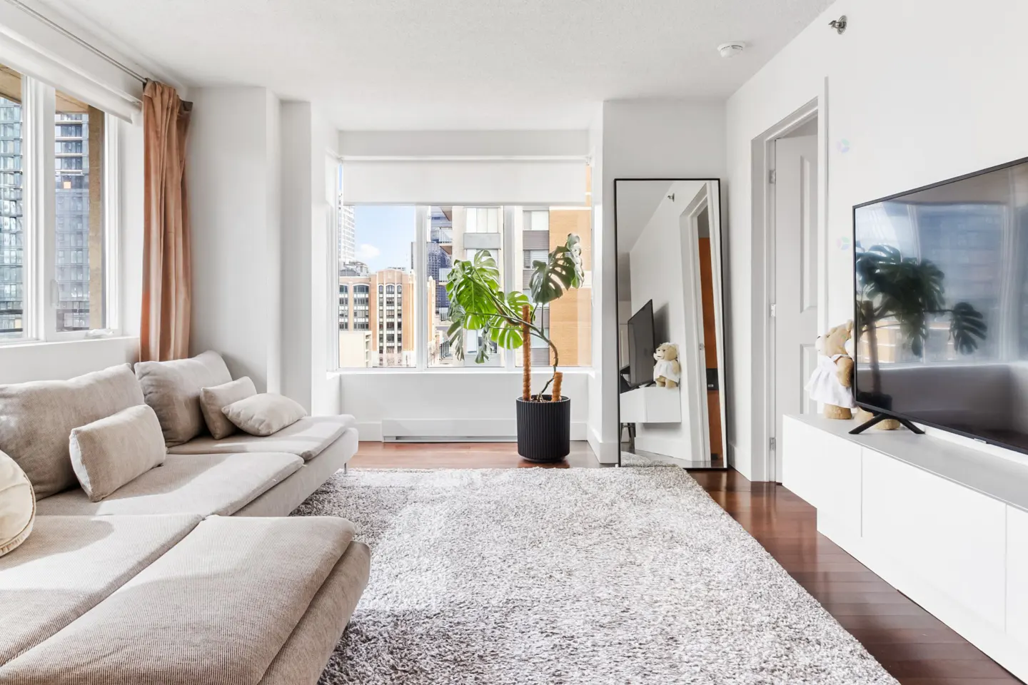 Bright living room with a beige sectional sofa, a large window with city views, and a black-framed mirror. A monstera plant sits near the window.
