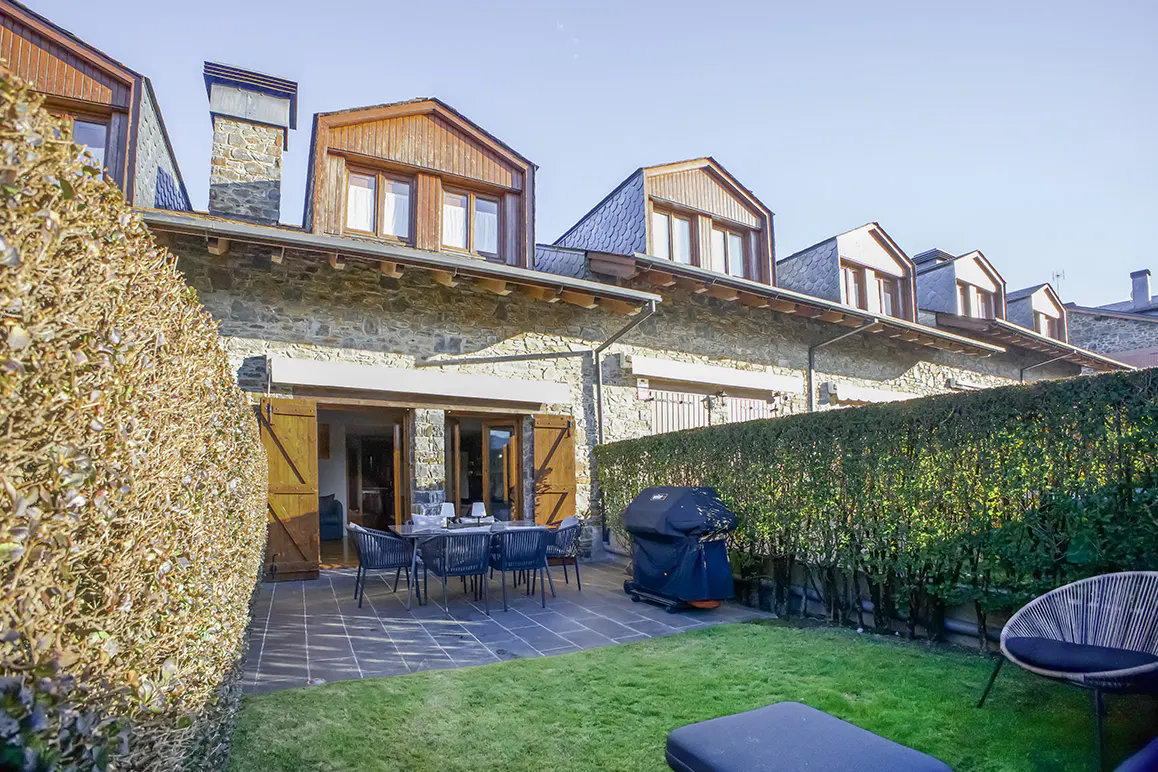 Exterior view of a stone house with a patio, table, chairs, grill, and green lawn.