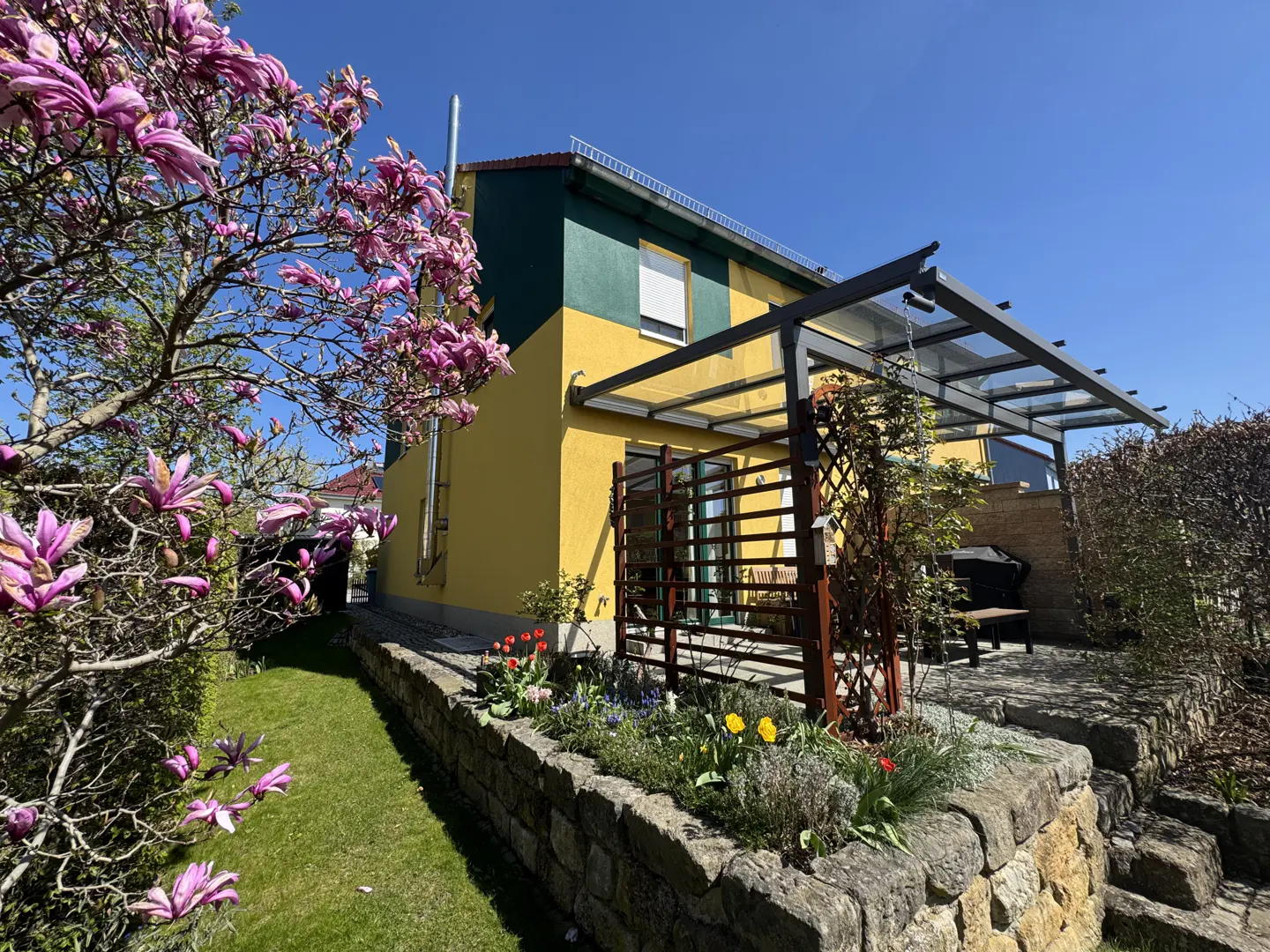Two-story house with yellow and green exterior, a patio with a pergola, and a stone garden with flowers.