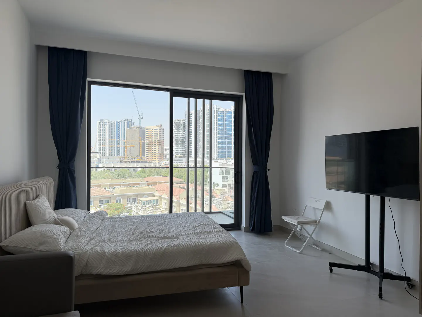 Modern bedroom with a bed, a wall-mounted TV on a stand, and a large window overlooking a city skyline with skyscrapers.