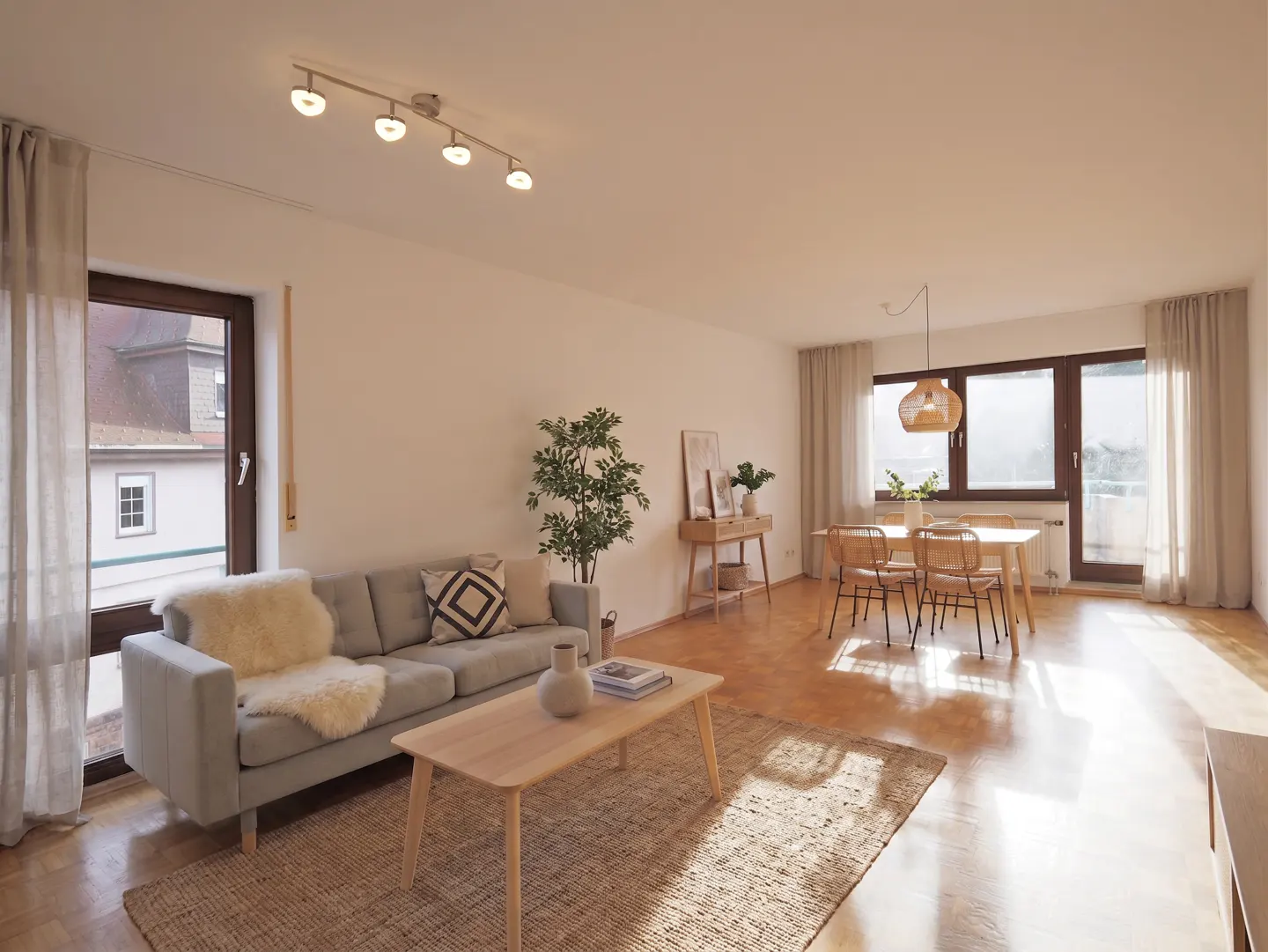 Bright, open-plan living and dining area with a grey sofa, wooden table, and dining set near large windows.