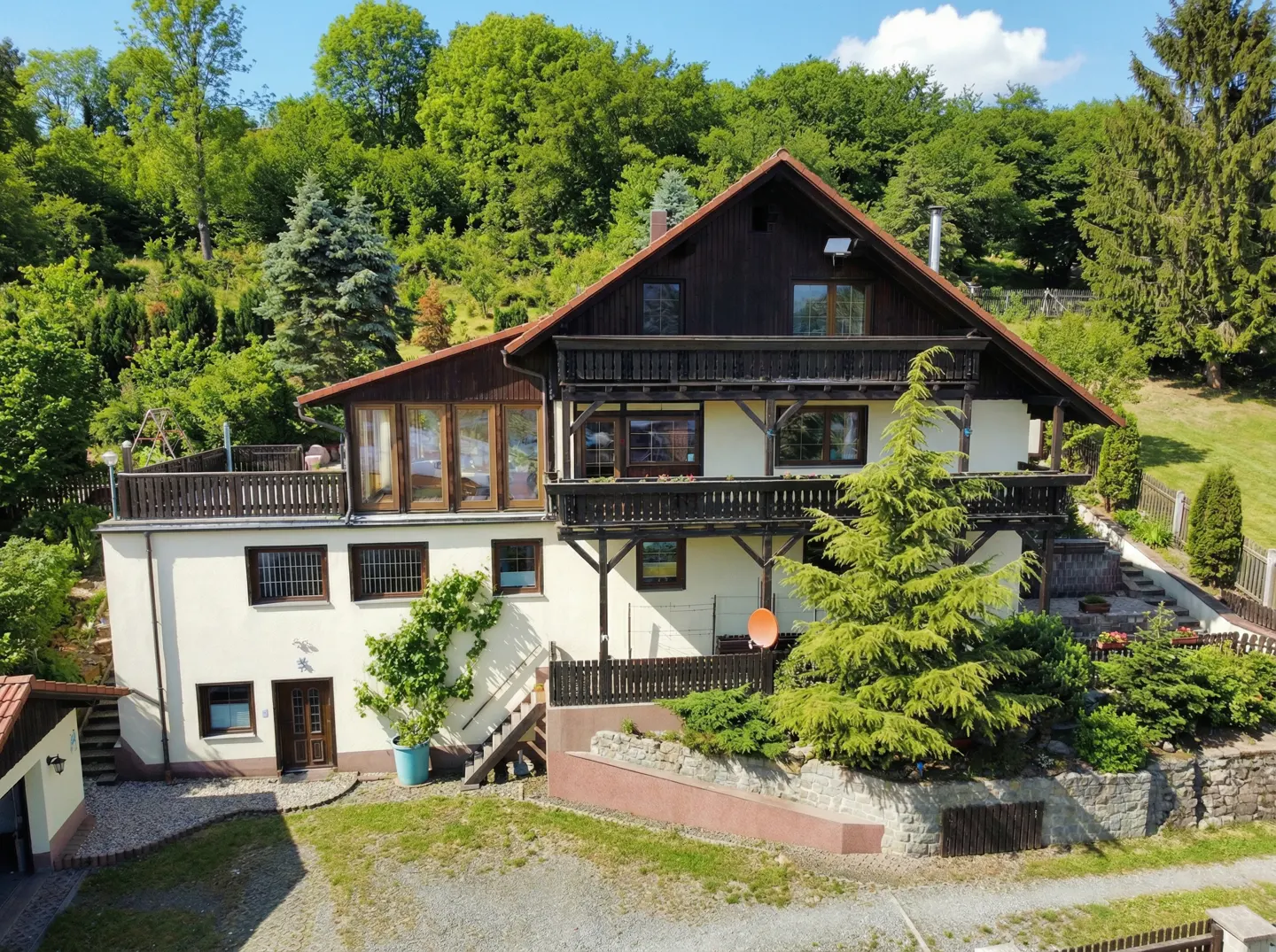 Exterior view of a two-story house with balconies, a sunroom, and a dark brown roof, surrounded by lush green trees.