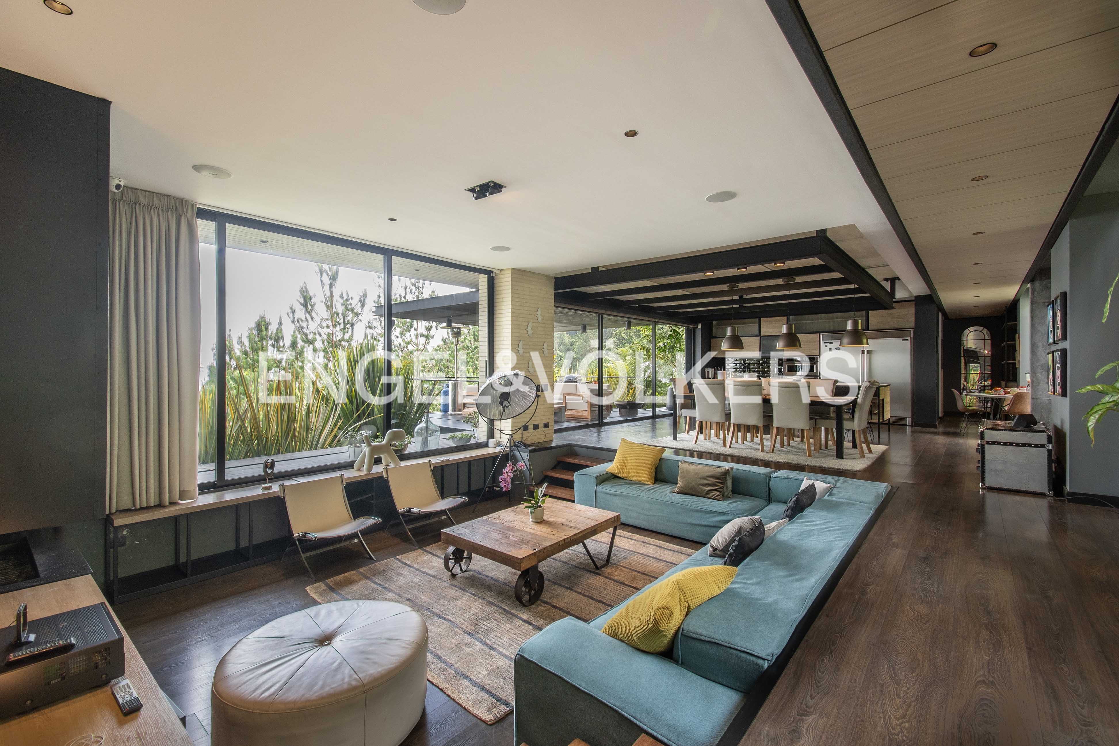 A spacious living room with a blue sofa, wooden floor, and large windows overlooking greenery. A dining area is visible in the background. [AI-generated text]