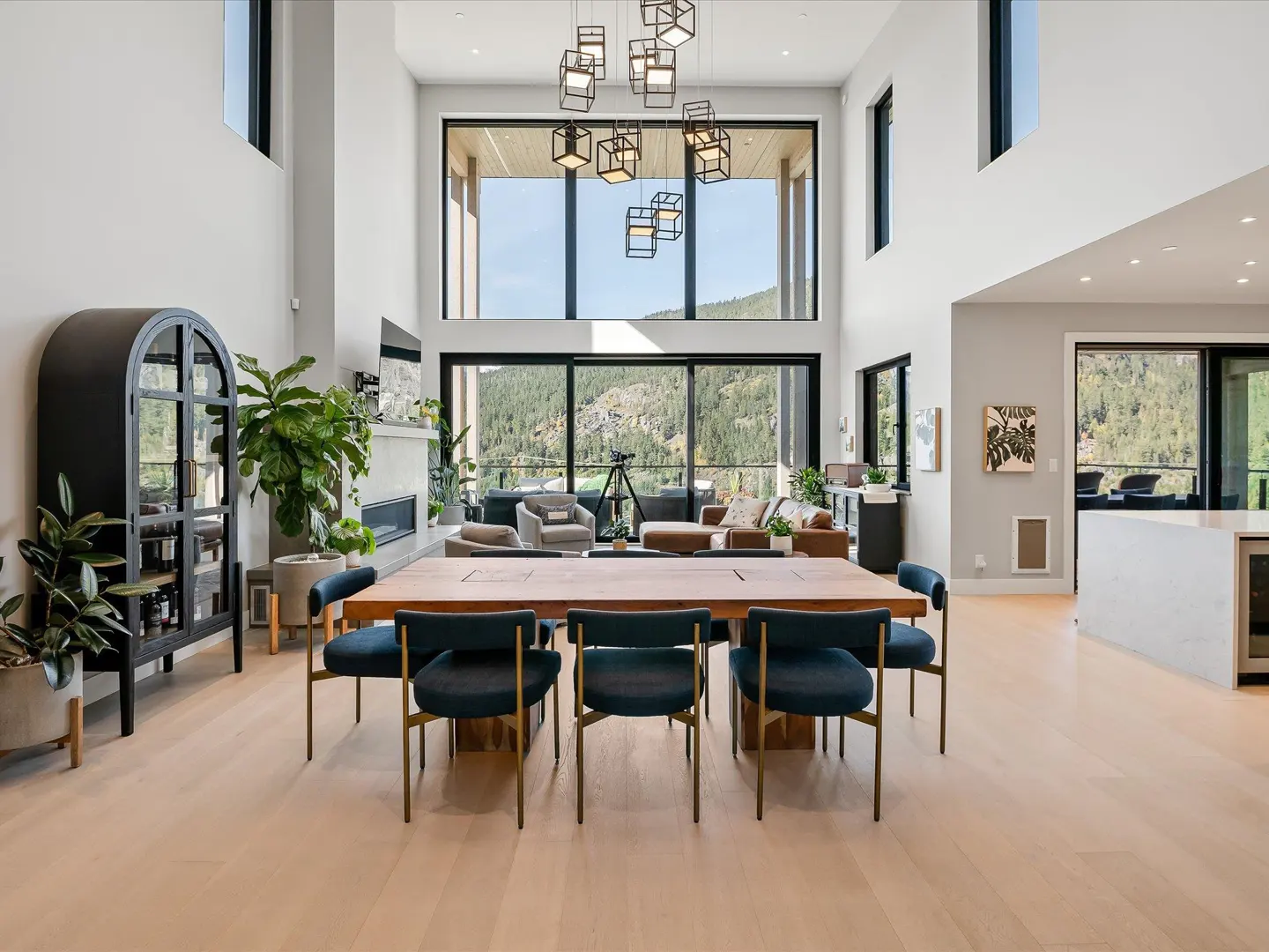 Bright, modern dining room with a large wooden table and blue chairs. Floor-to-ceiling windows offer mountain views. Cube chandelier hangs above.