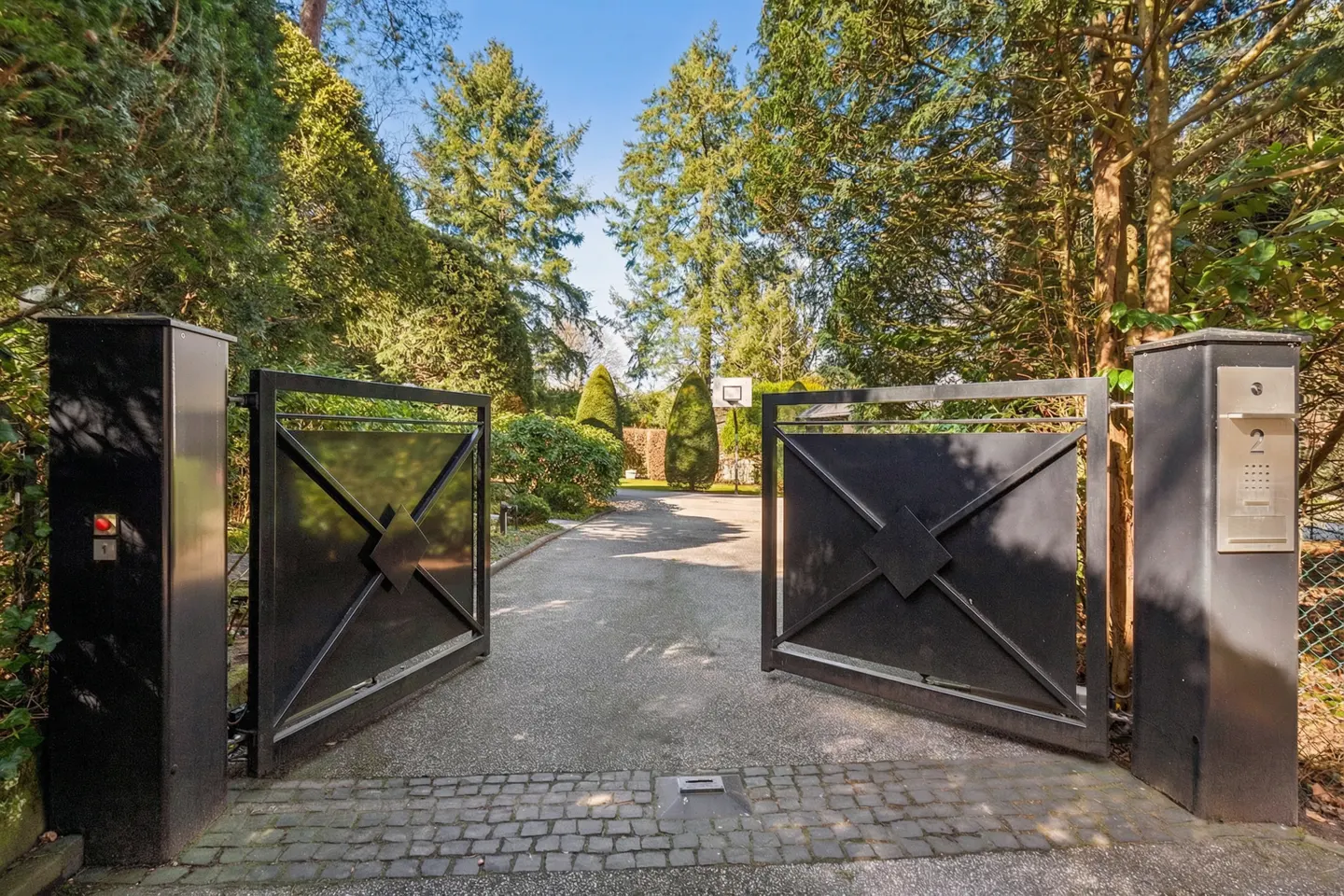 Open black metal gate leading to a driveway, with trees and a basketball hoop visible in the background.