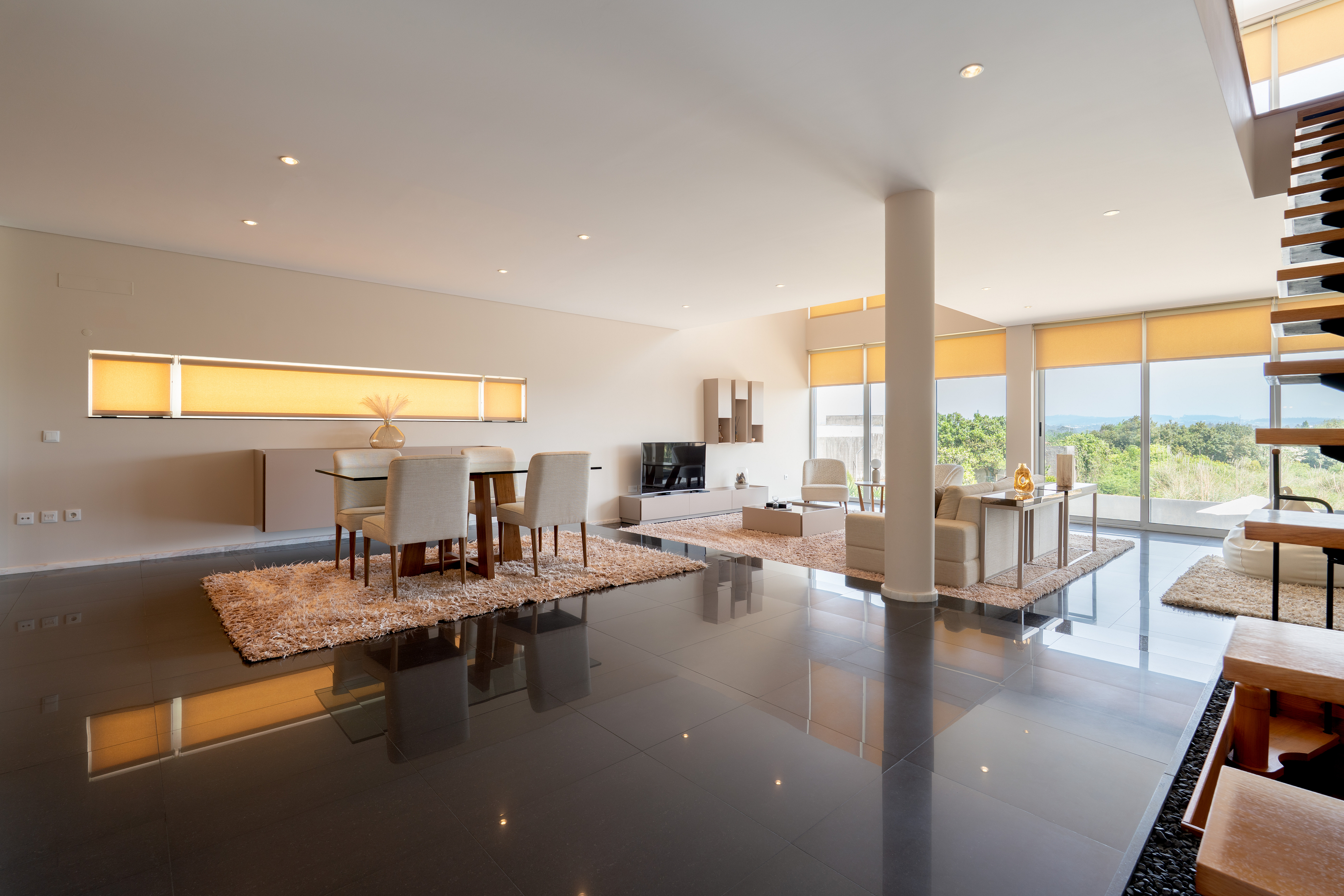 Open-concept living space with dining table, chairs, and living room furniture. Large windows with yellow shades overlook greenery.