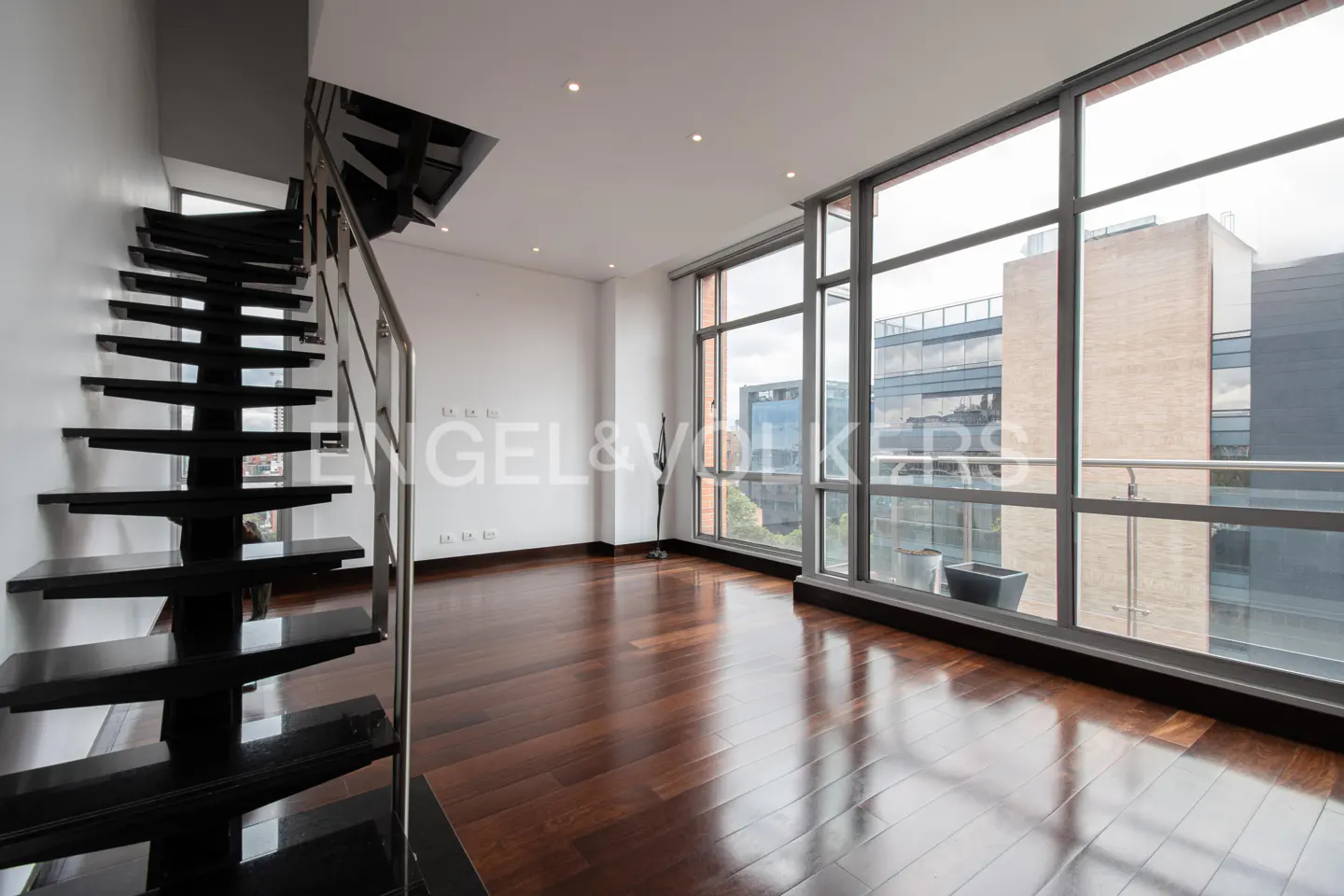 Bright, modern apartment interior with dark wood floors, black stairs, and large windows overlooking city buildings.