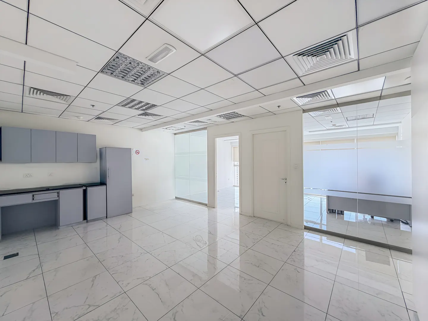 Bright, empty office space with white tile floors, gray cabinets, and glass-walled rooms.