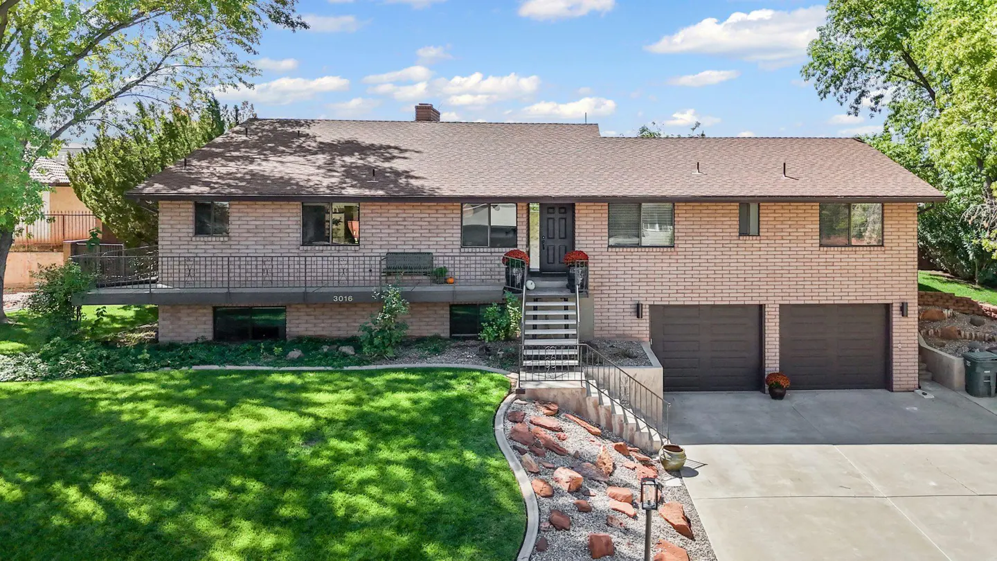 Two-story brick house with a brown roof, dark trim, and a two-car garage. A green lawn and trees surround the house.
