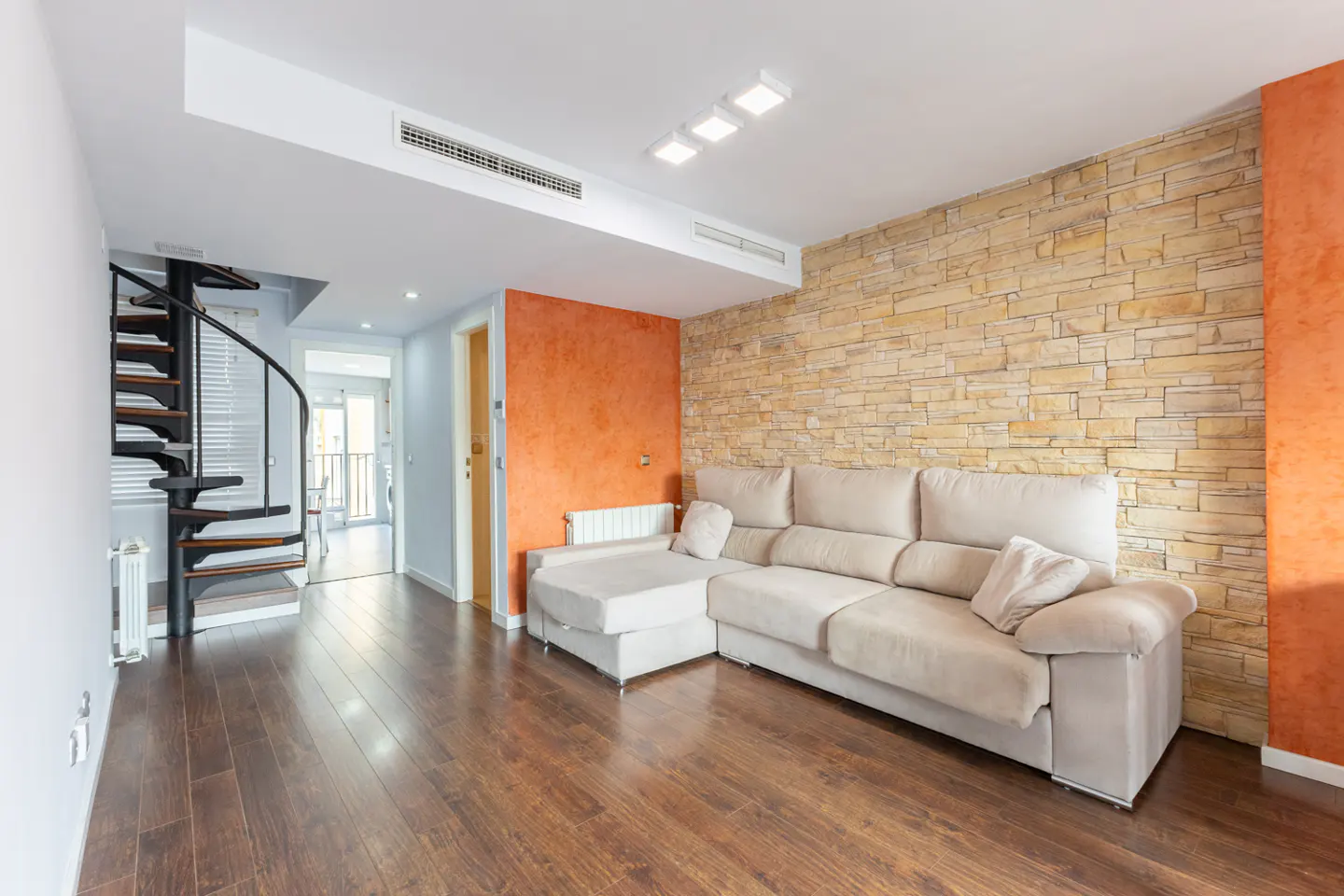 Living room with a beige sectional sofa, orange accent wall, and stone wall. A black spiral staircase is visible in the background.