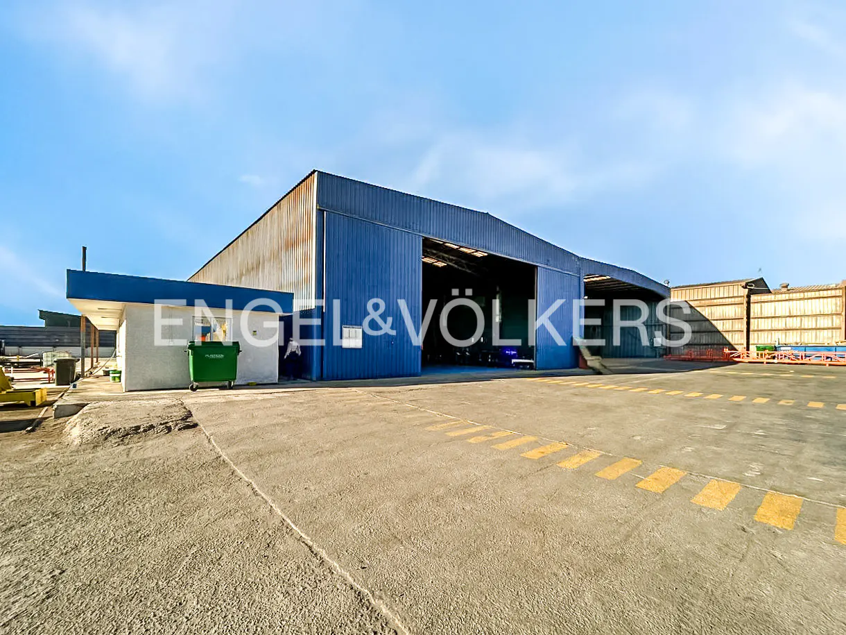 Large blue industrial warehouse with an open bay, a white side office, and a green dumpster on a paved lot under blue sky.