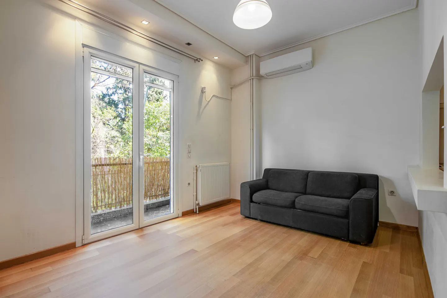 Bright, empty room with wood floors, white walls, and a dark gray sofa. Balcony with bamboo screen visible through glass doors.
