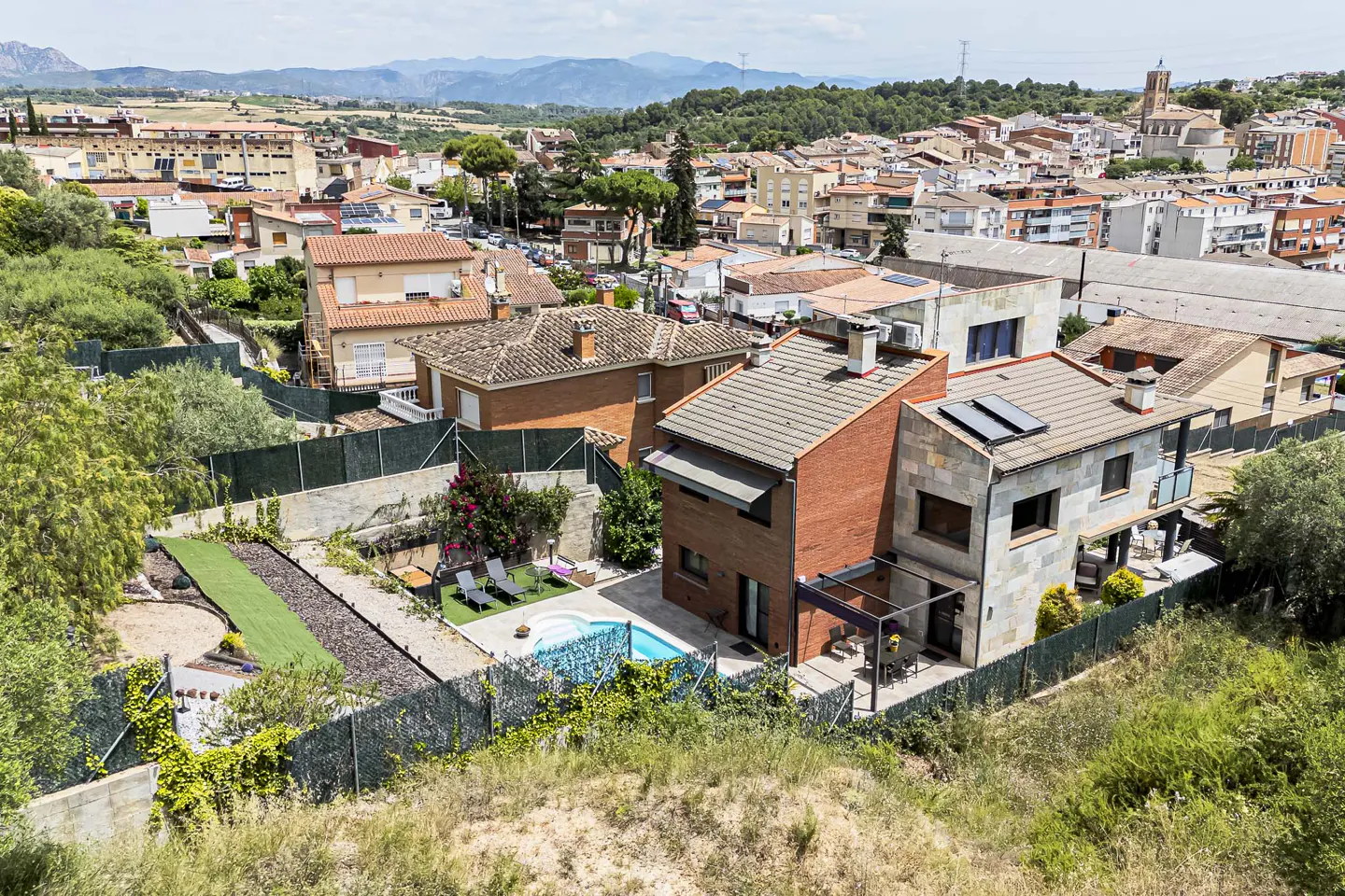 Aerial view of a modern brick and stone house with a backyard pool, patio, and lush garden overlooking a Spanish town.