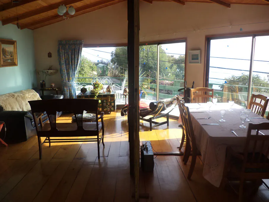 Bright living and dining room with wood floors and ceiling. Large windows overlook a green landscape and ocean view. Table set for a meal.
