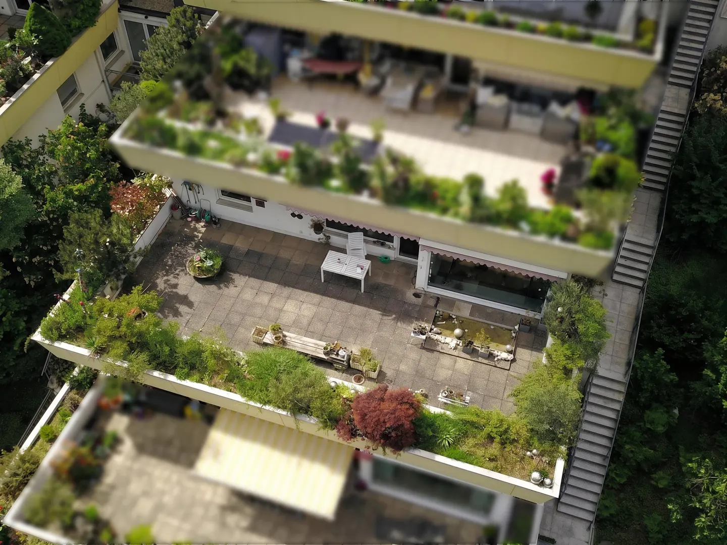 Aerial view of tiered apartments with rooftop gardens and patios, connected by outdoor stairs. Lush greenery and outdoor furniture are visible.