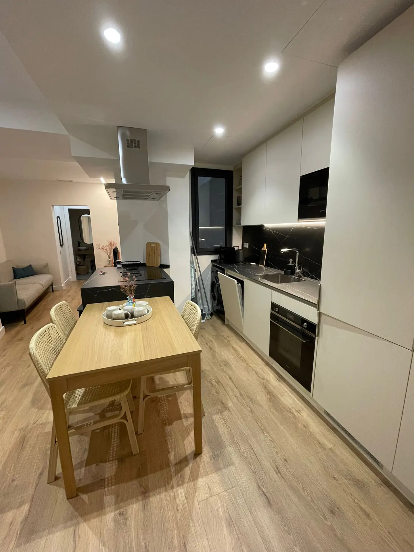 Modern kitchen with white cabinets, light wood dining table, and chairs, featuring a black island and wood-look flooring.