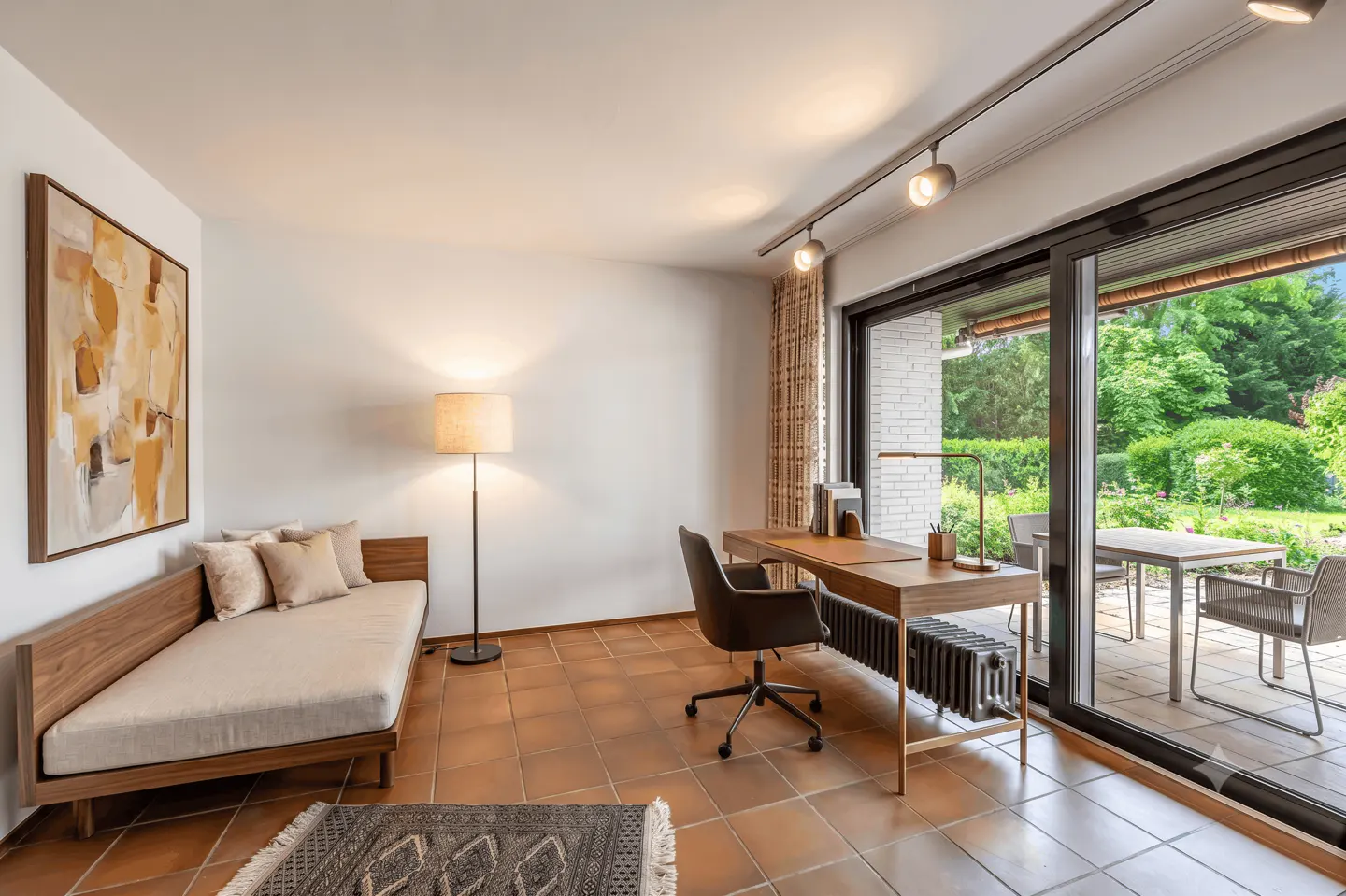 A bright room with terracotta tile floors features a daybed, desk, and sliding glass doors to a patio and garden.