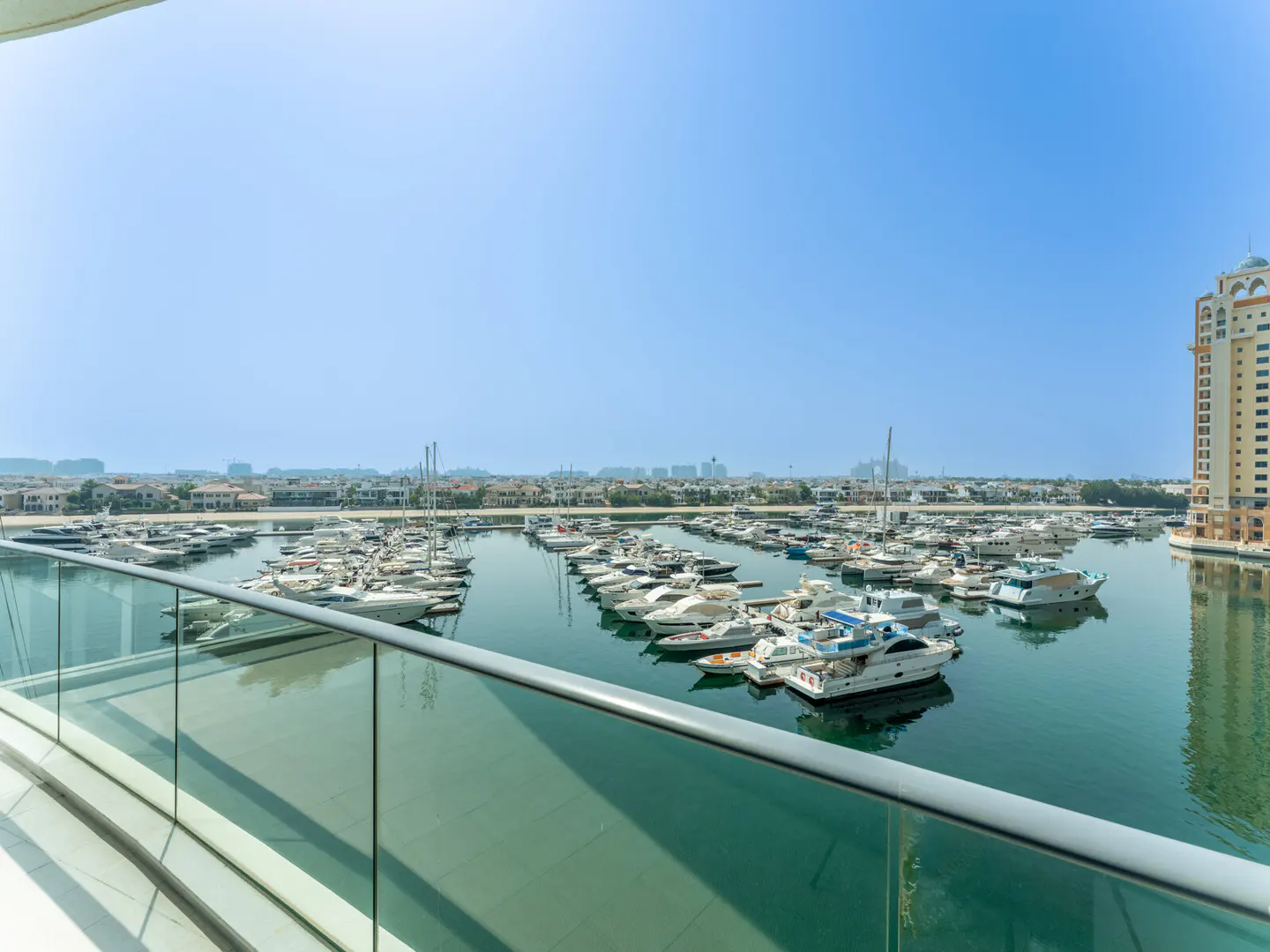 View from a balcony overlooking a marina filled with yachts on a sunny day. A tall building is visible on the right.