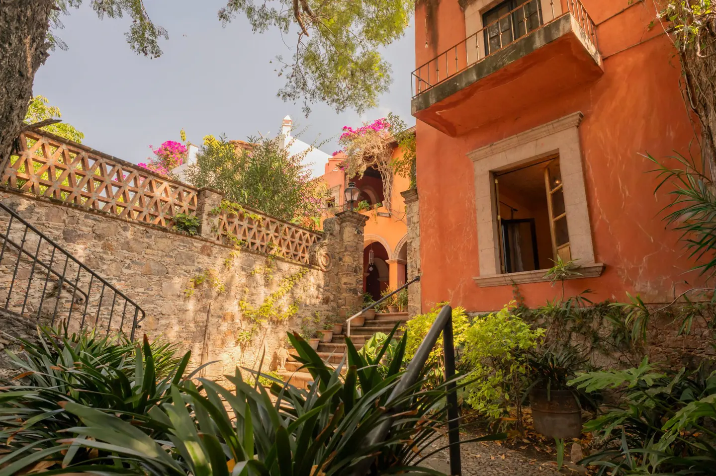 Exterior view of an orange building with a balcony, stone wall, stairs, and lush greenery.
