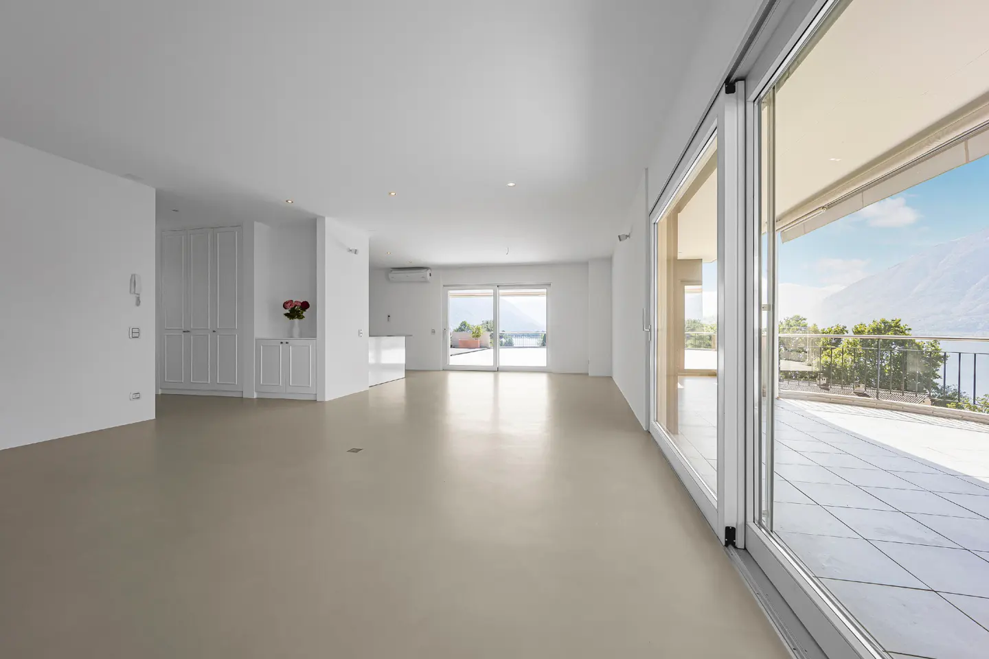 Bright, empty room with white walls, beige floor, and sliding glass doors to a balcony with a view of a lake and mountains.