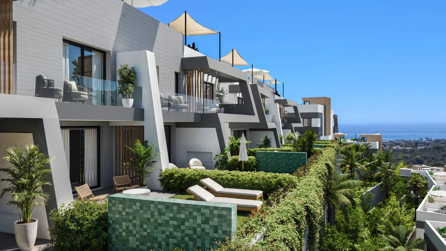 Modern grey and white apartments with balconies and rooftop terraces overlook the ocean. Greenery and lounge chairs add to the luxurious feel.