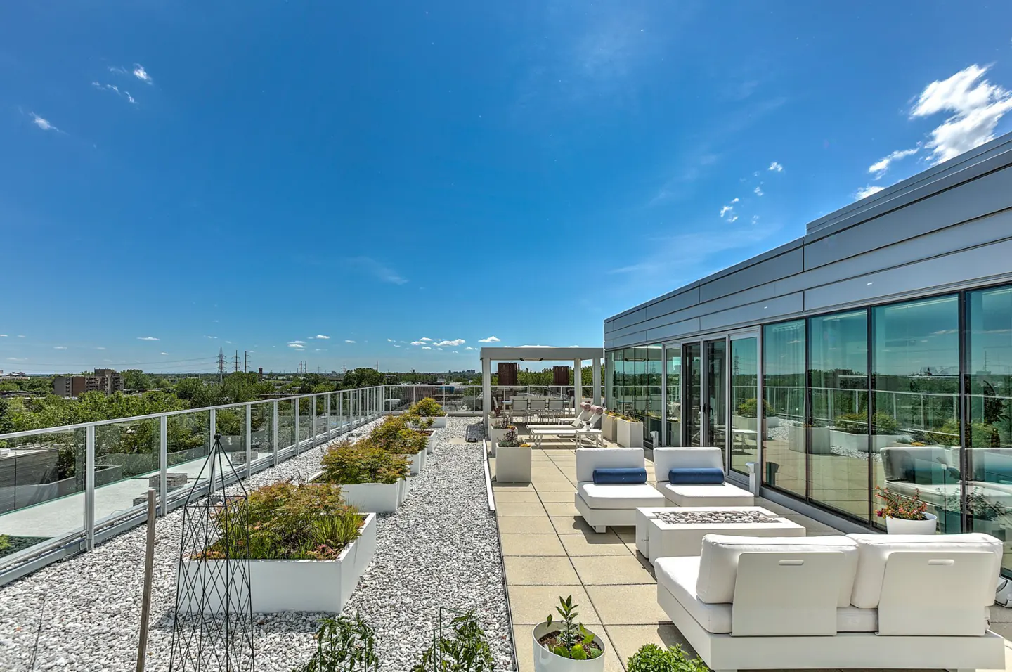 Rooftop patio with white lounge furniture, planters, and gravel. Glass railing overlooks green trees under a blue sky.