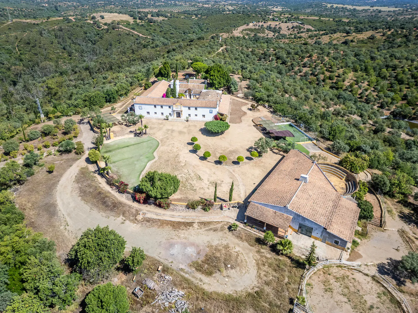 Aerial view of a large white house with a red tile roof, surrounded by green trees and brown fields.