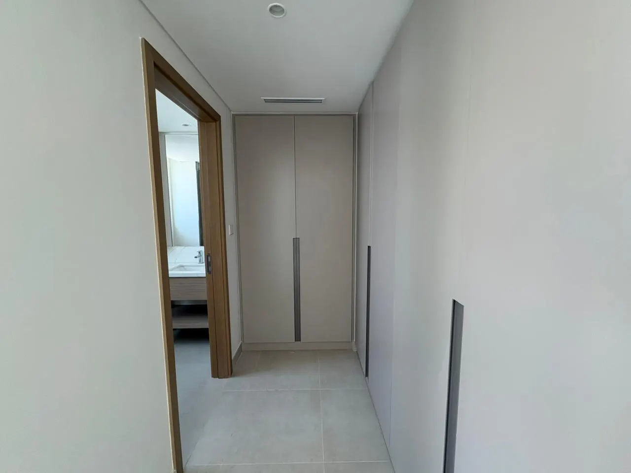 Hallway with light gray walls, tile floor, and a built-in closet. A doorway leads to a bathroom with a sink and mirror.