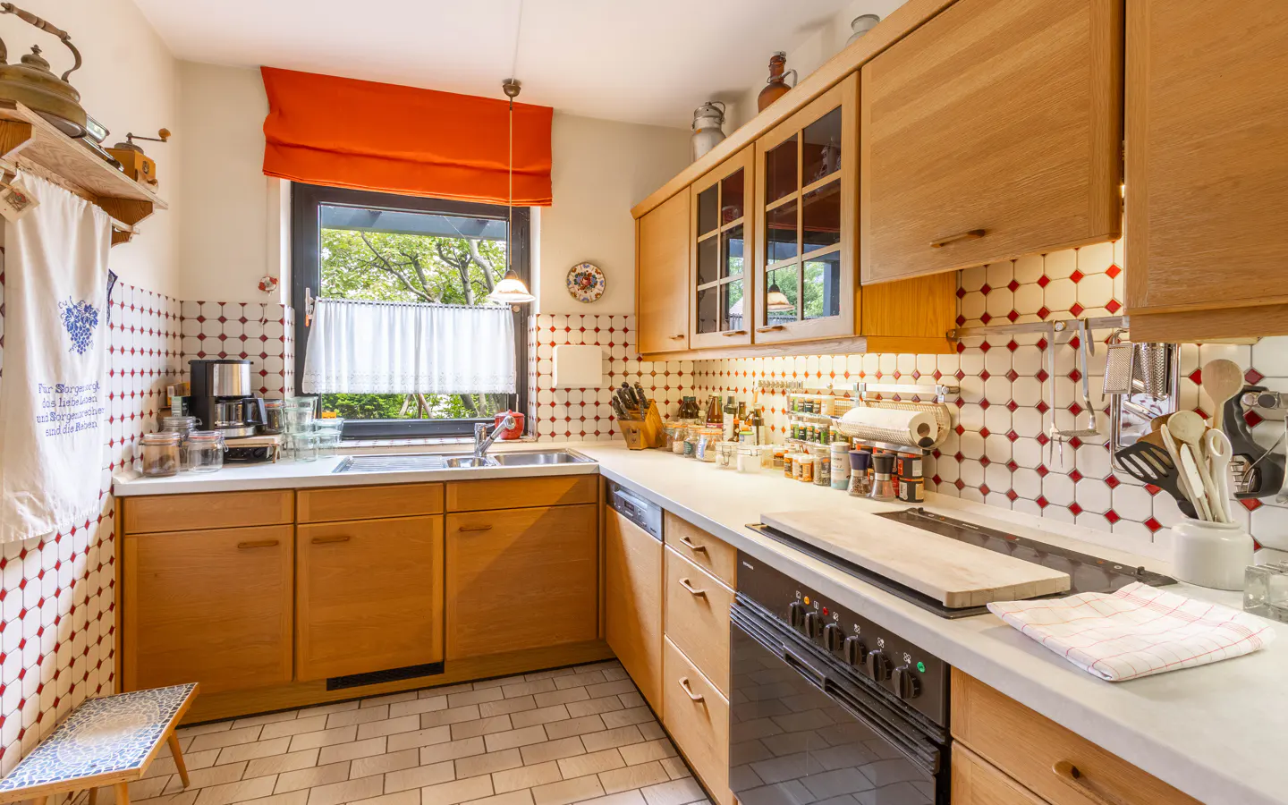 Cozy kitchen with light wood cabinets, white countertops, and a red-and-white diamond tile backsplash under a bright window.