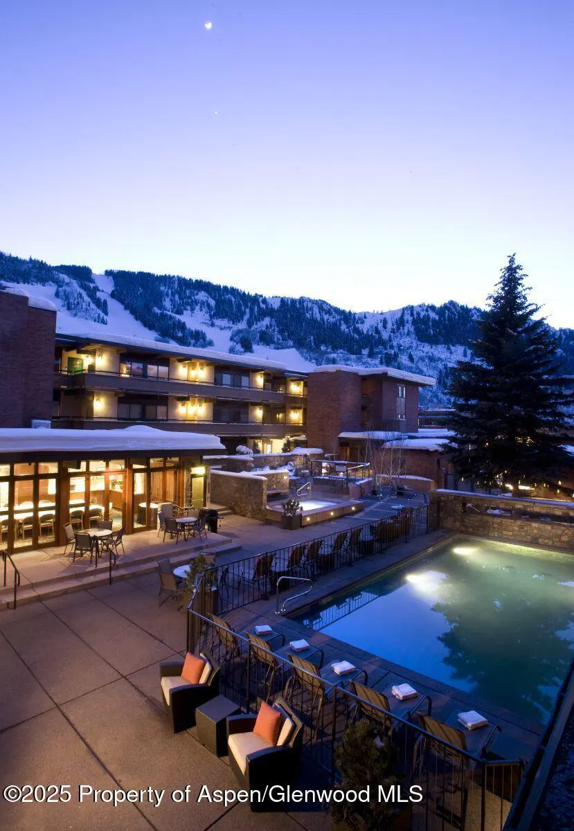 Exterior view of a resort with a pool, lounge chairs, and snow-covered mountains in the background at dusk.