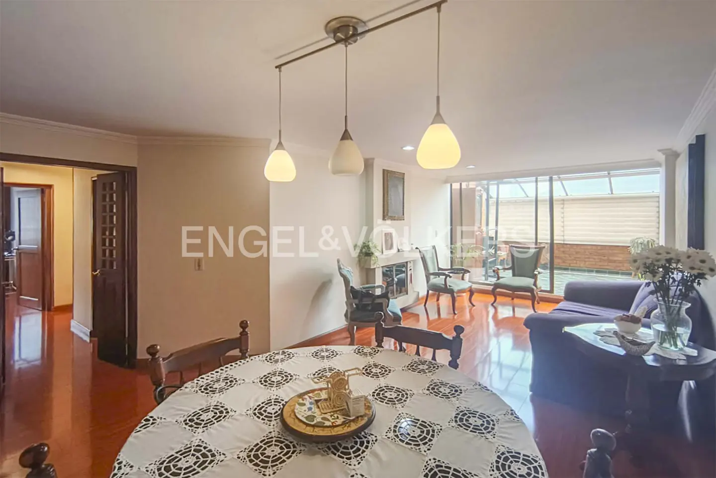 A bright living and dining area with hardwood floors, a round table with a white tablecloth, and a seating area with a blue sofa. Three pendant lights hang above the table.