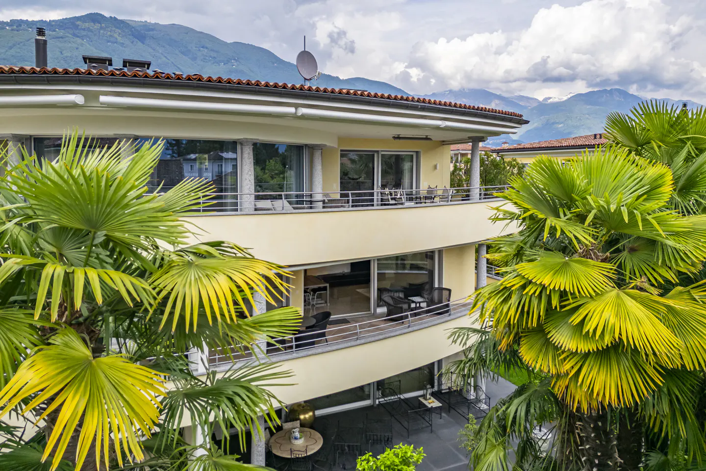Two-story yellow house with balconies, surrounded by palm trees. Mountains are visible in the background.
