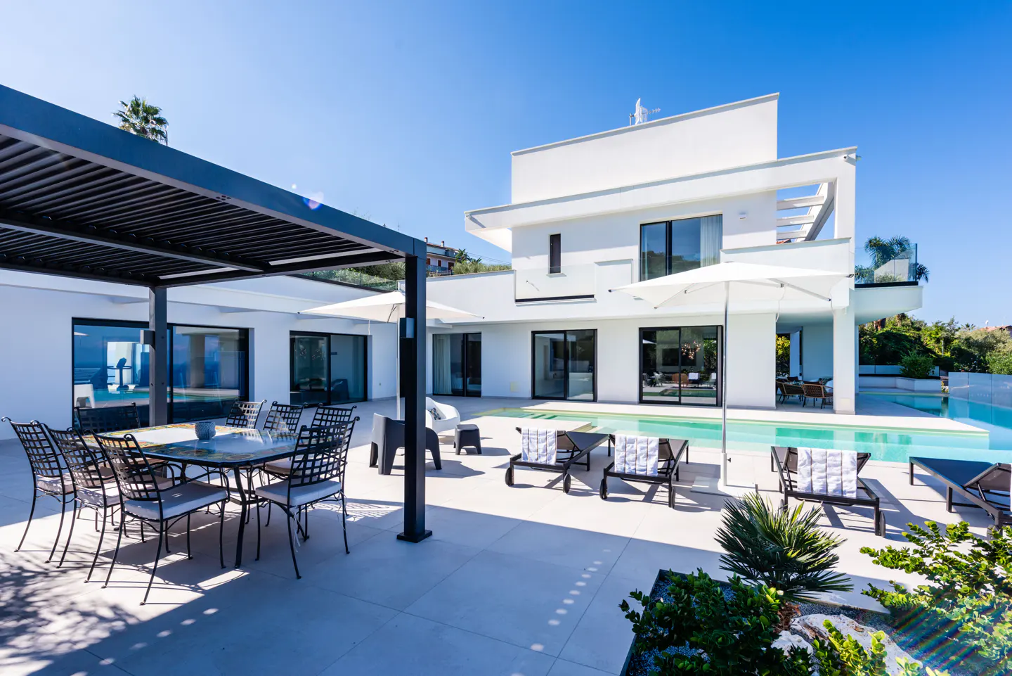 Modern white house with a pool, lounge chairs, and outdoor dining area under a black pergola on a sunny day.