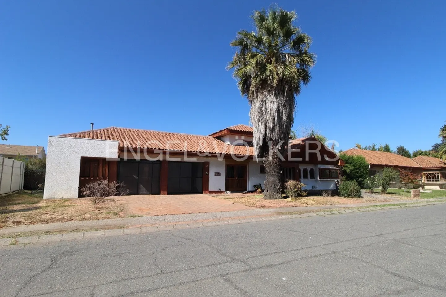 A white stucco house with a red tile roof and a two-car garage under a clear blue sky. A tall palm tree stands in the front yard.
