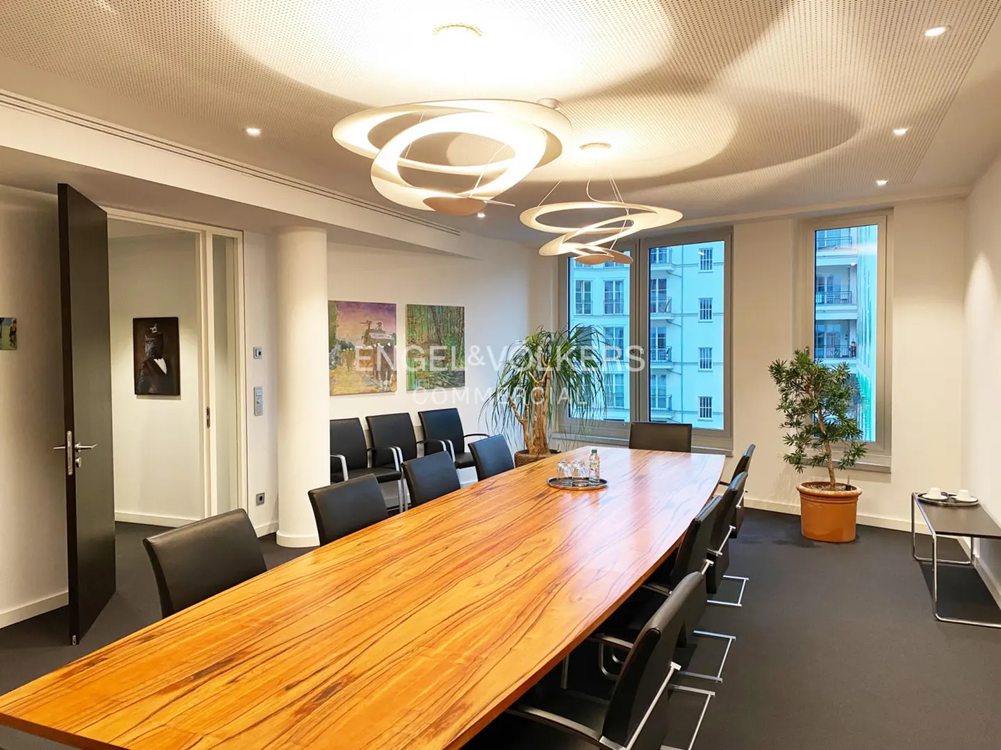 Modern conference room with a long wooden table, black chairs, artistic wall paintings, and unique circular light fixtures.