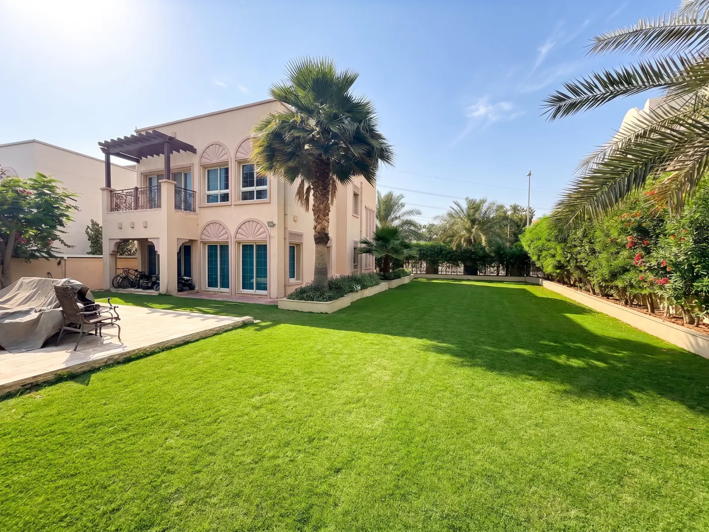 Two-story beige house with a large green lawn, palm trees, and a blue sky.